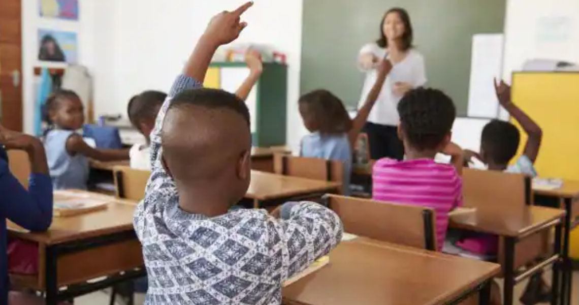 Students raising their hands in a classroom, as the teacher points to one of them