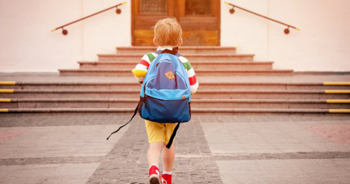 Rear view of a young boy wearing a backpack and walking toward a school building