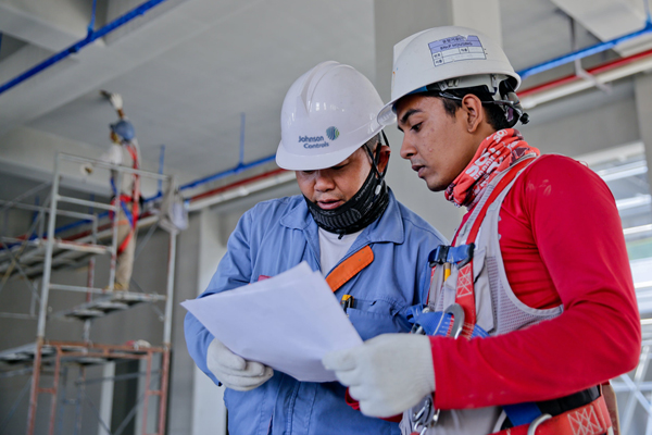 Two maintenance workers wearing Johnson Controls helmets, looking at a chart in a facility