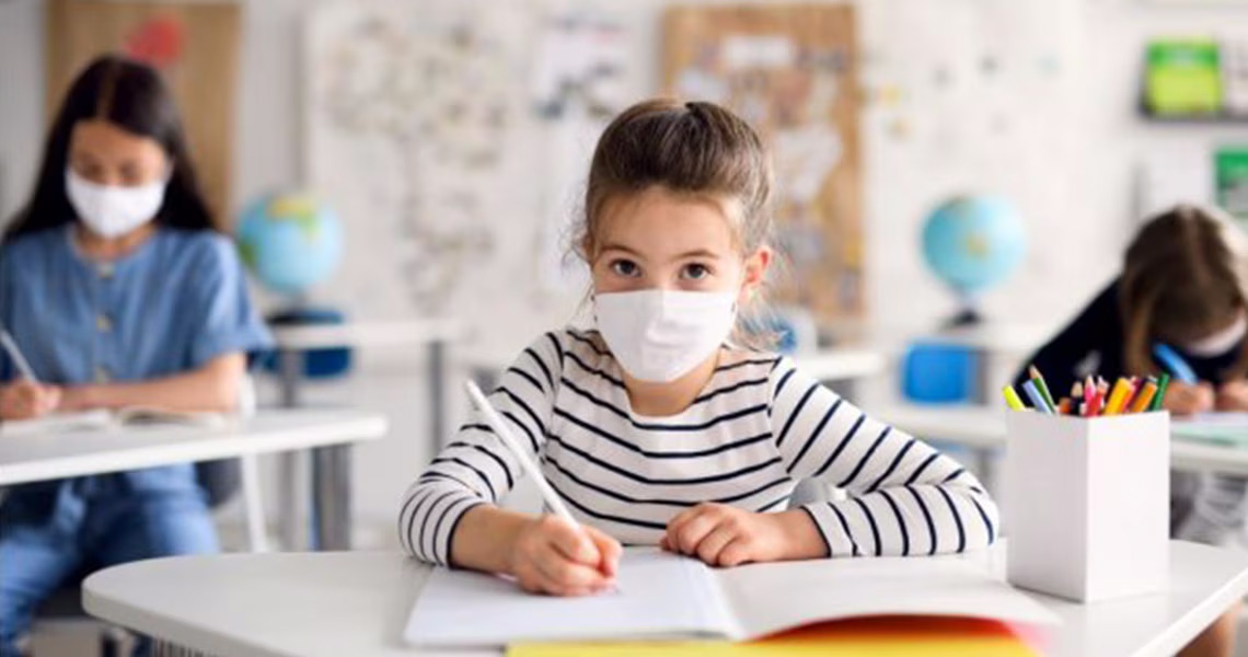 A young girl seated at a desk in a classroom, wearing a facemask