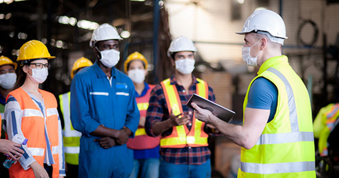 A diverse group of factory workers wearing white hardhats and masks, discussing work