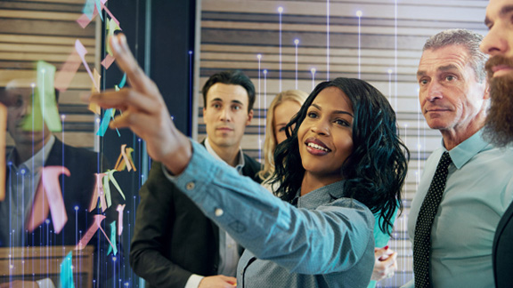 A woman pointing at sticky notes stuck on a glass wall in an office, while her collegaues watch